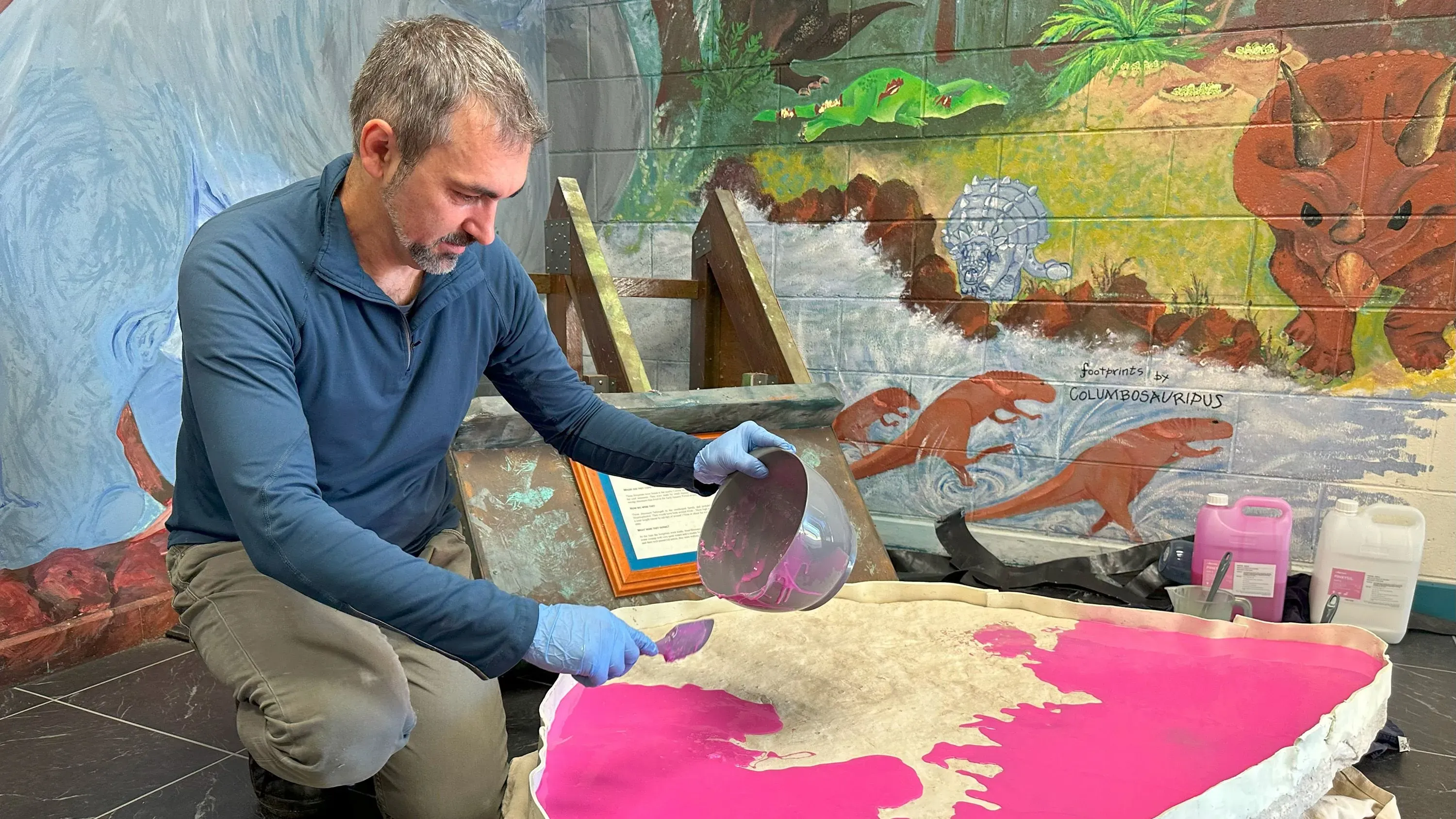 عالم حفريات يعمل على تحليل لوح حجري يحتوي على آثار أقدام ديناصورات من العصر الجوراسي المبكر، مع خلفية فنية توضح الديناصورات.