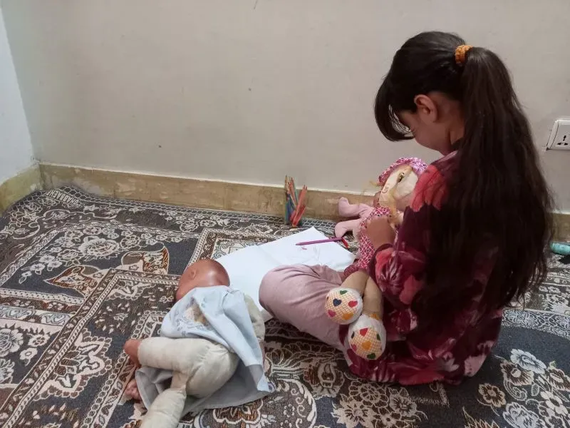 طفلة أفغانية تلعب بدمية وتكتب على ورقة في غرفة بسيطة، تعكس تأثير الأحداث المروعة على حياتها اليومية.