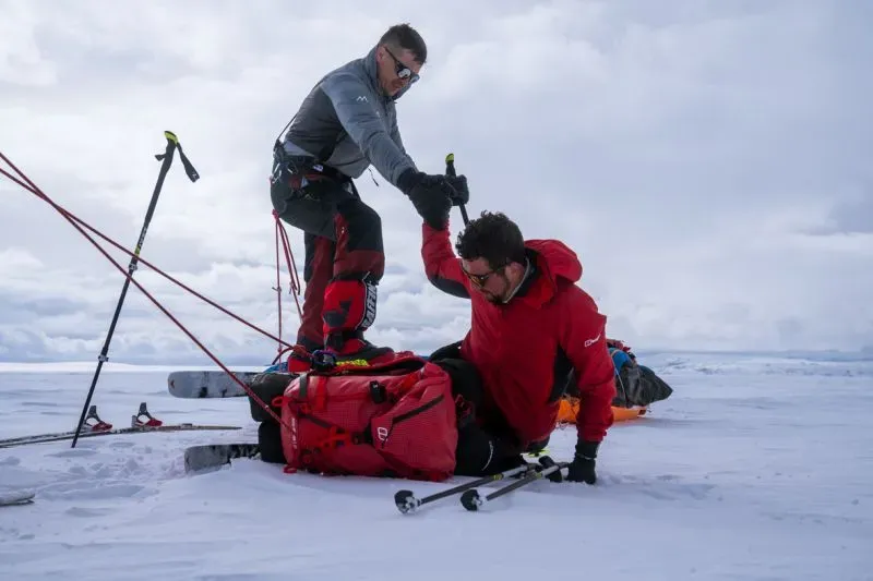 صورة تظهر دارين إدواردز يتلقى المساعدة من صديقه أثناء التزلج في بيئة ثلجية، مع معدات التزلج في الخلفية.