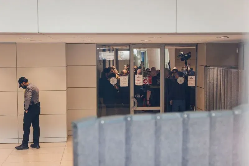 موظف حكومي يقف بجوار باب زجاجي، بينما يجتمع عدد من الأشخاص في الداخل، مما يعكس القلق بشأن المراقبة في ظل إدارة ترامب.