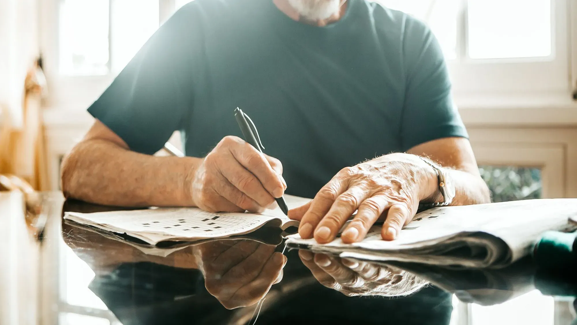 رجل مسن يحل لغز الكلمات المتقاطعة على طاولة، مما يعكس أهمية الأنشطة العقلية النشطة في الحفاظ على صحة الدماغ.