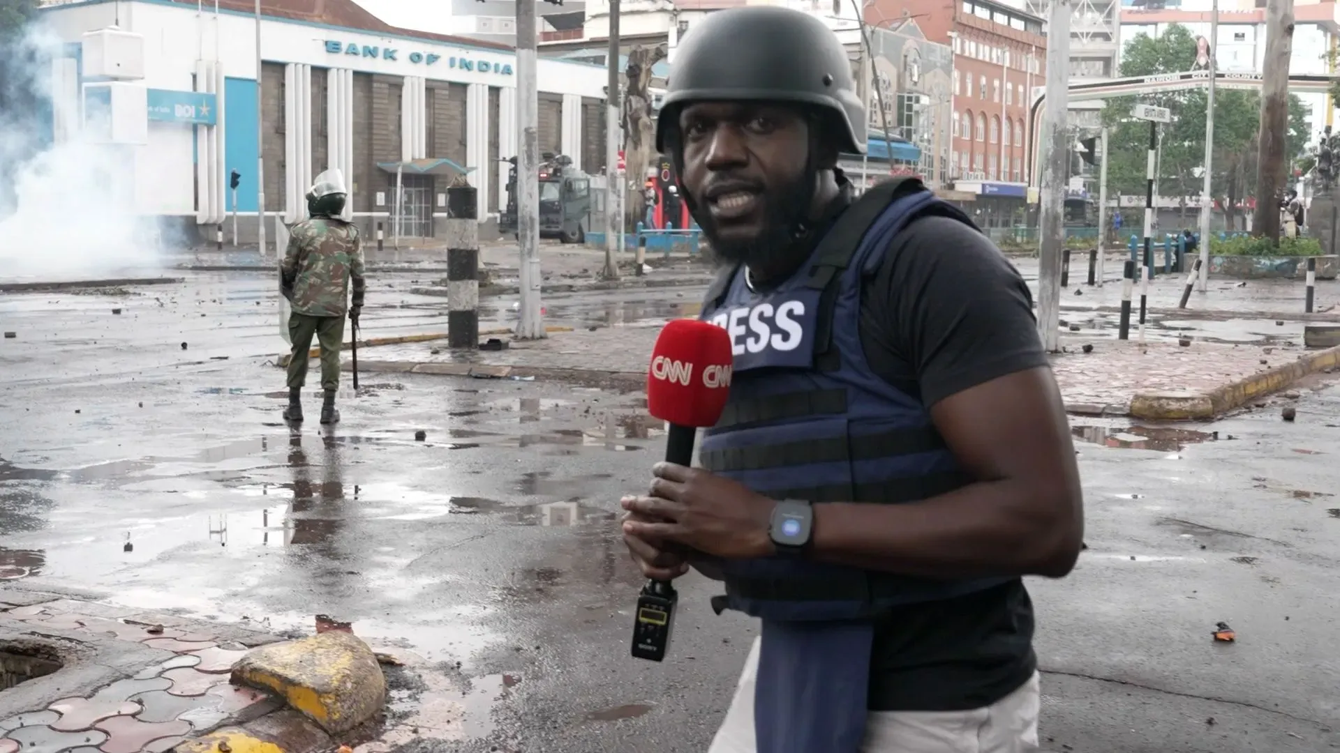 صحفي يرتدي درع واقٍ وخوذة، يقف في موقع مظاهرة في نيروبي، بينما تظهر آثار الفوضى والغاز المسيل للدموع في الخلفية.