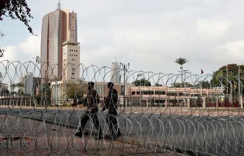 شخصان يسيران بجوار أسلاك شائكة تحيط بمبنى البرلمان الكيني في نيروبي، وسط أجواء التوتر قبل المظاهرات ضد الحكومة.