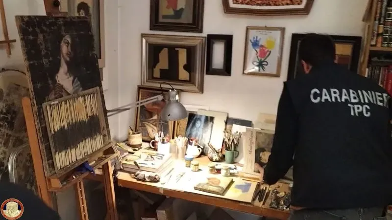 ورشة فنية في روما تحتوي على لوحات مزيفة وأدوات رسم، بينما يقوم أحد رجال الشرطة بتفتيش المكان بحثًا عن الأدلة.