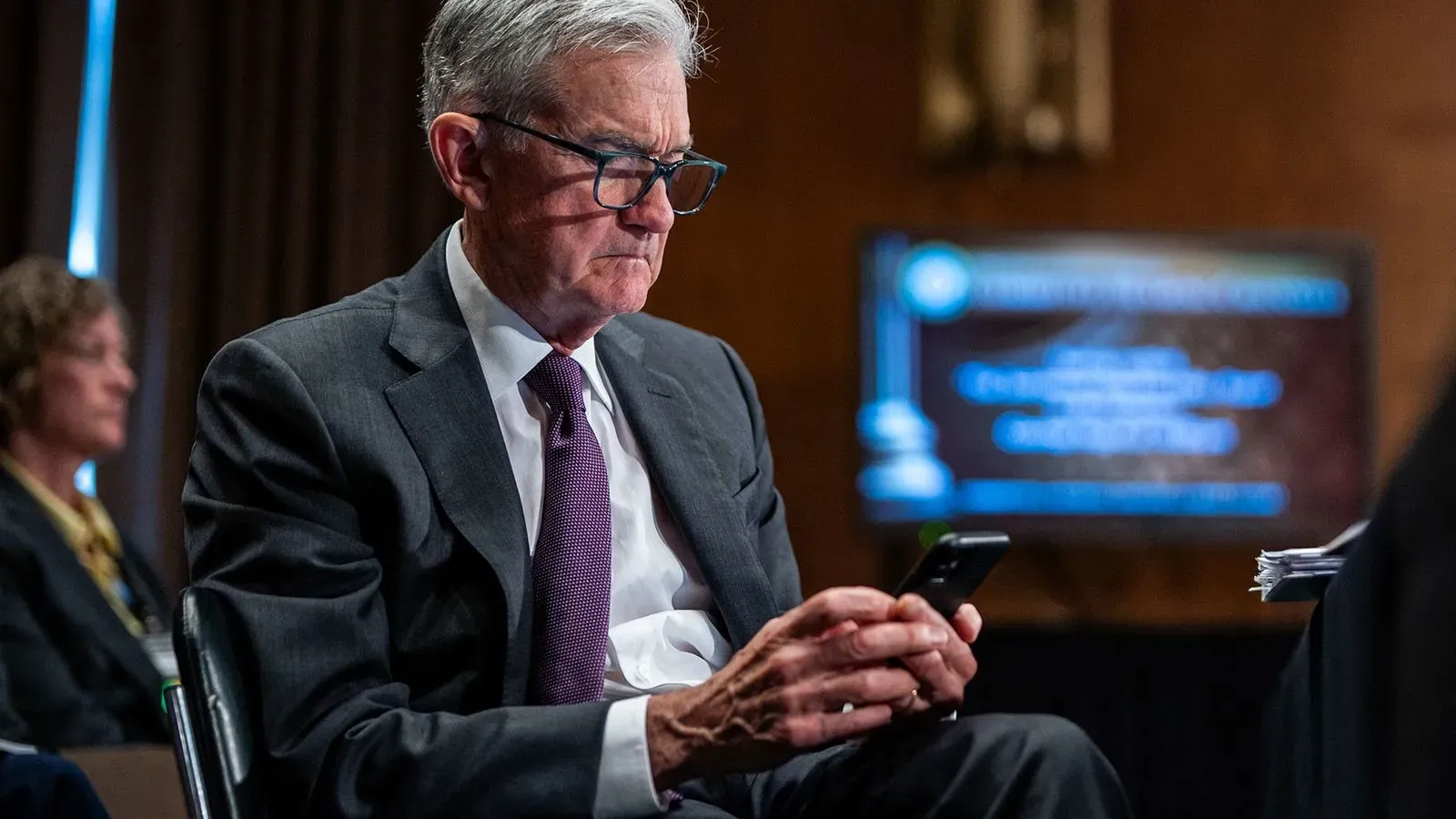 جيروم باول، رئيس مجلس الاحتياطي الفيدرالي، يتصفح هاتفه خلال جلسة استماع، مع وجود شاشة عرض خلفه تتعلق بالسياسة النقدية.