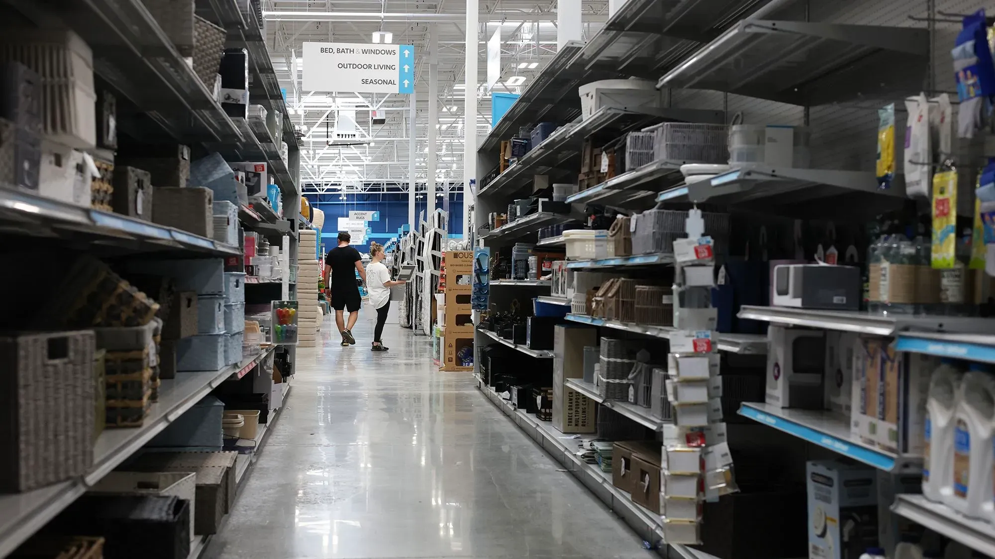 ممر في متجر يعرض رفوفًا فارغة جزئيًا، مع زبائن يتجولون، مما يعكس تأثير الرسوم الجمركية على توفر السلع.