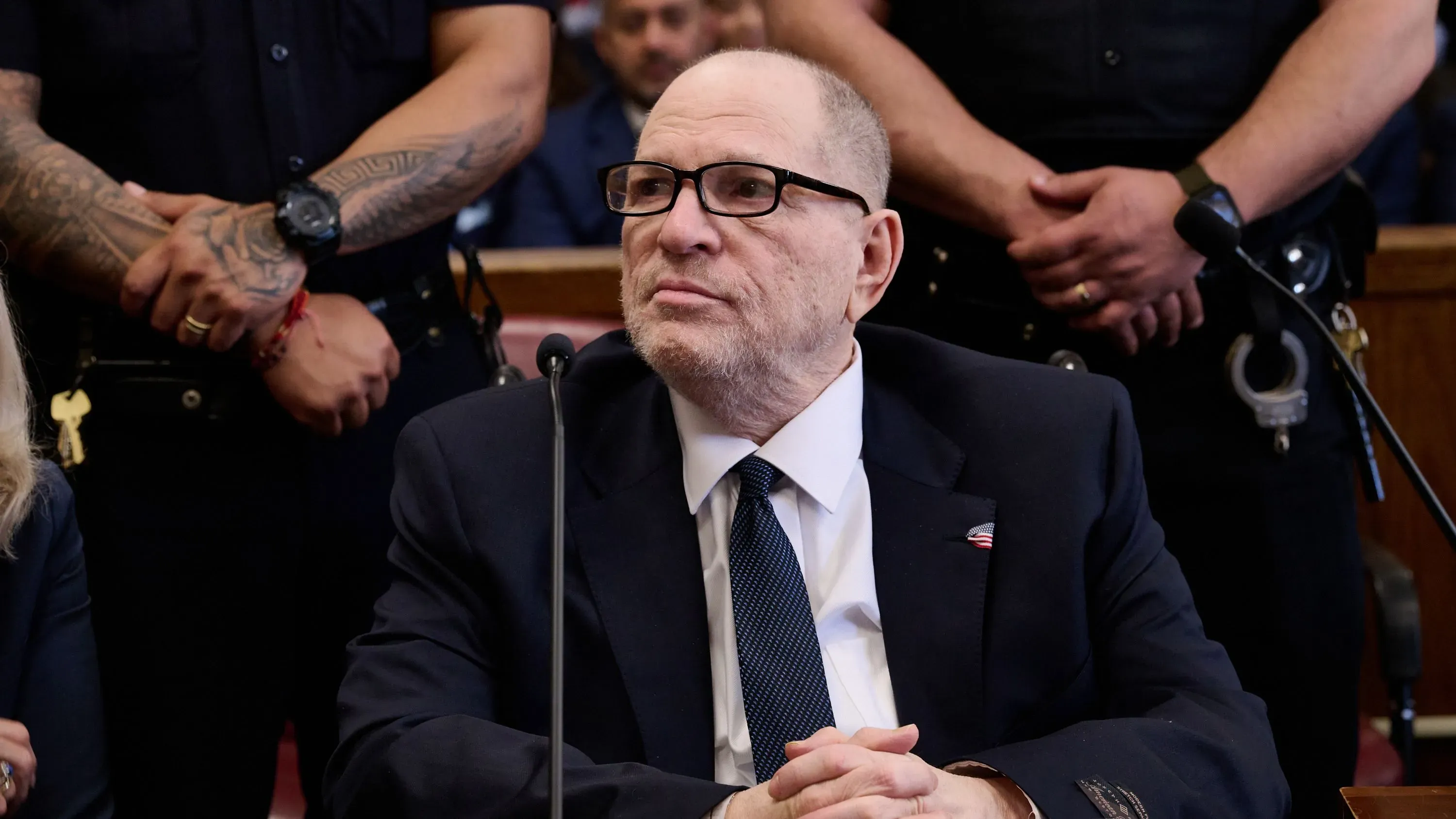 هارفي واينستين يجلس في قاعة المحكمة أثناء محاكمته بتهم الاعتداء الجنسي، محاطًا بأفراد من الشرطة.