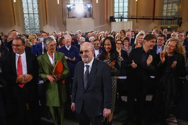 سلمان رشدي، المؤلف المعروف، يقف وسط حشد من الناس في حدث رسمي، مرتديًا نظارات شمسية، مع تعبيرات تعكس مشاعر مختلطة من الدعم والاهتمام.