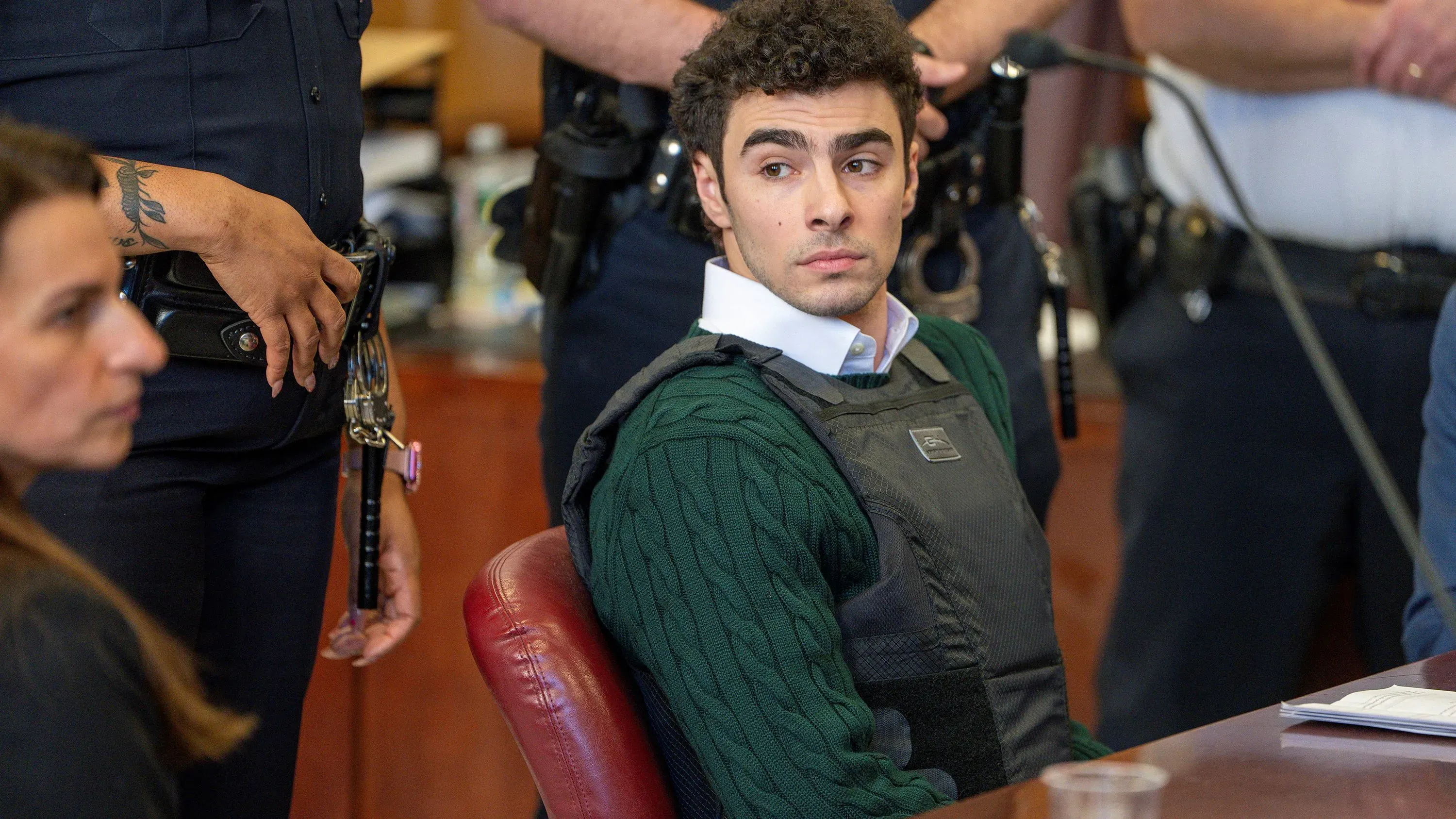 لويجي مانجيوني في قاعة المحكمة، مرتديًا سترة واقية من الرصاص وقميصًا أخضر، محاطًا بأفراد من الشرطة، وسط أجواء مشحونة.