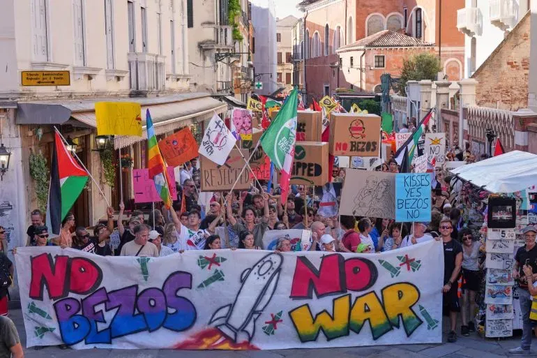 محتجون في شوارع البندقية يحملون لافتات تعبر عن رفضهم لزفاف جيف بيزوس، مع شعارات مثل "لا بيزوس، لا حرب" و"قبلات نعم، بيزوس لا".