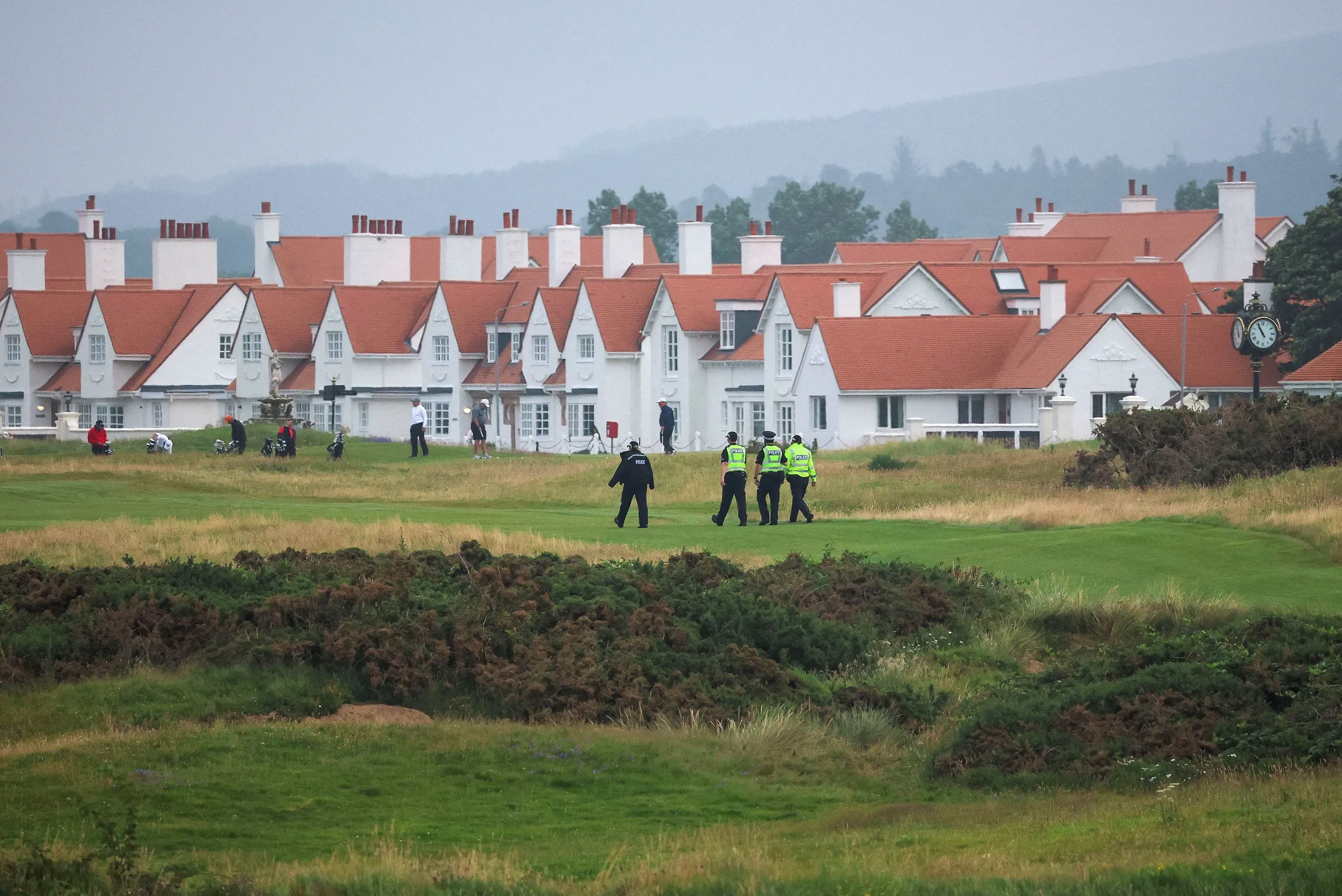تظهر الصورة مجموعة من رجال الشرطة يسيرون على ملعب غولف في اسكتلندا، مع منازل ذات أسطح حمراء خلفهم، في ظل أجواء ضبابية.