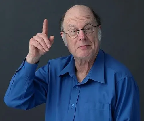 عالم فيزياء الكم تشارلز بينيت يرتدي قميصًا أزرق، يشير بإصبعه لأعلى، مع تعبير وجه يعكس التفكير والابتكار في مجال التشفير الكمي.