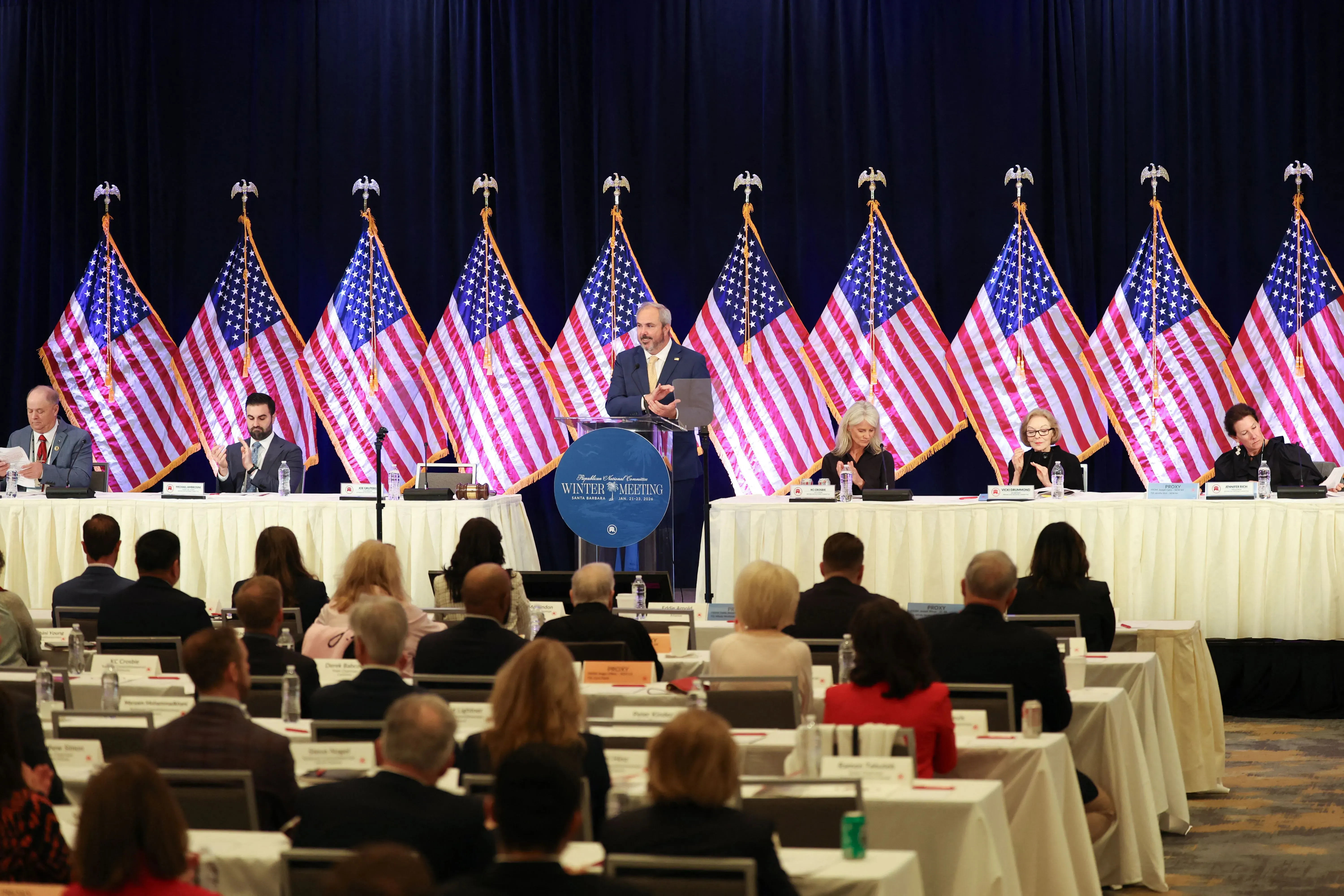 اجتماع اللجنة الوطنية للحزب الجمهوري في سانتا باربرا، حيث يتحدث أحد القادة أمام طاولة مغطاة بأعلام الولايات المتحدة، وسط حضور كبير.