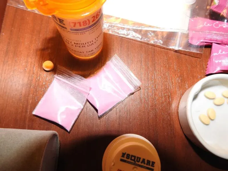 زجاجة دواء تحمل اسم "فرانك بلاك" مع حبوب وعبوات تحتوي على مسحوق وردي، يُعتقد أنه كيتامين، على طاولة في غرفة نوم كومبس.