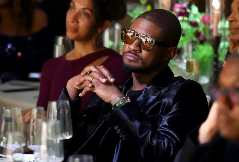 صورة لمغني الراب "أشر" مرتديًا نظارات شمسية، يجلس في حدث اجتماعي مع تعبير جاد، بينما تظهر امرأة خلفه. تعكس الصورة أجواء الموسيقى والثقافة.