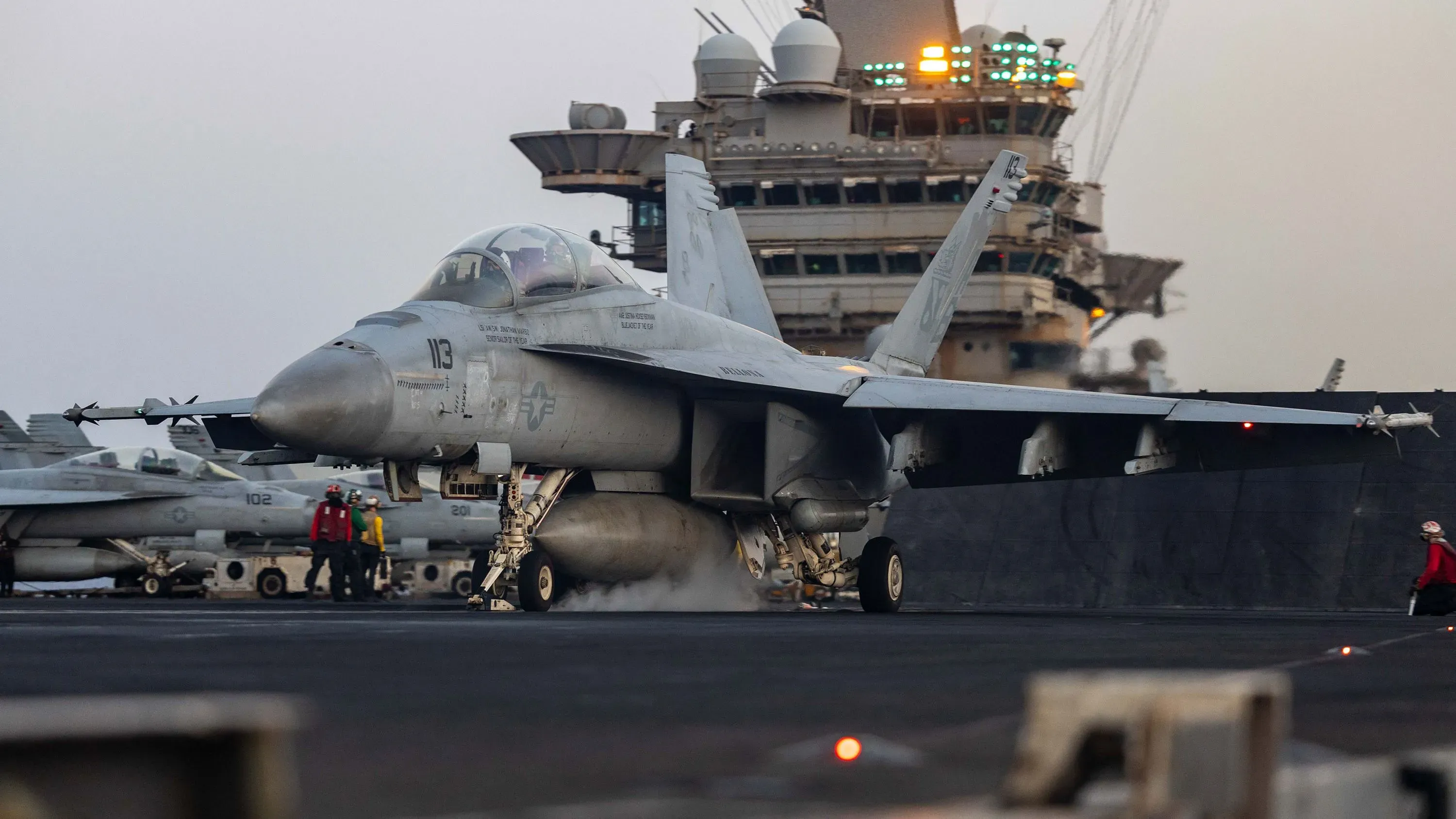 طائرة F/A-18 سوبر هورنيت على متن حاملة الطائرات هاري ترومان، مع طاقم يعمل في الخلفية، في سياق حوادث الطائرات الأخيرة في البحر الأحمر.