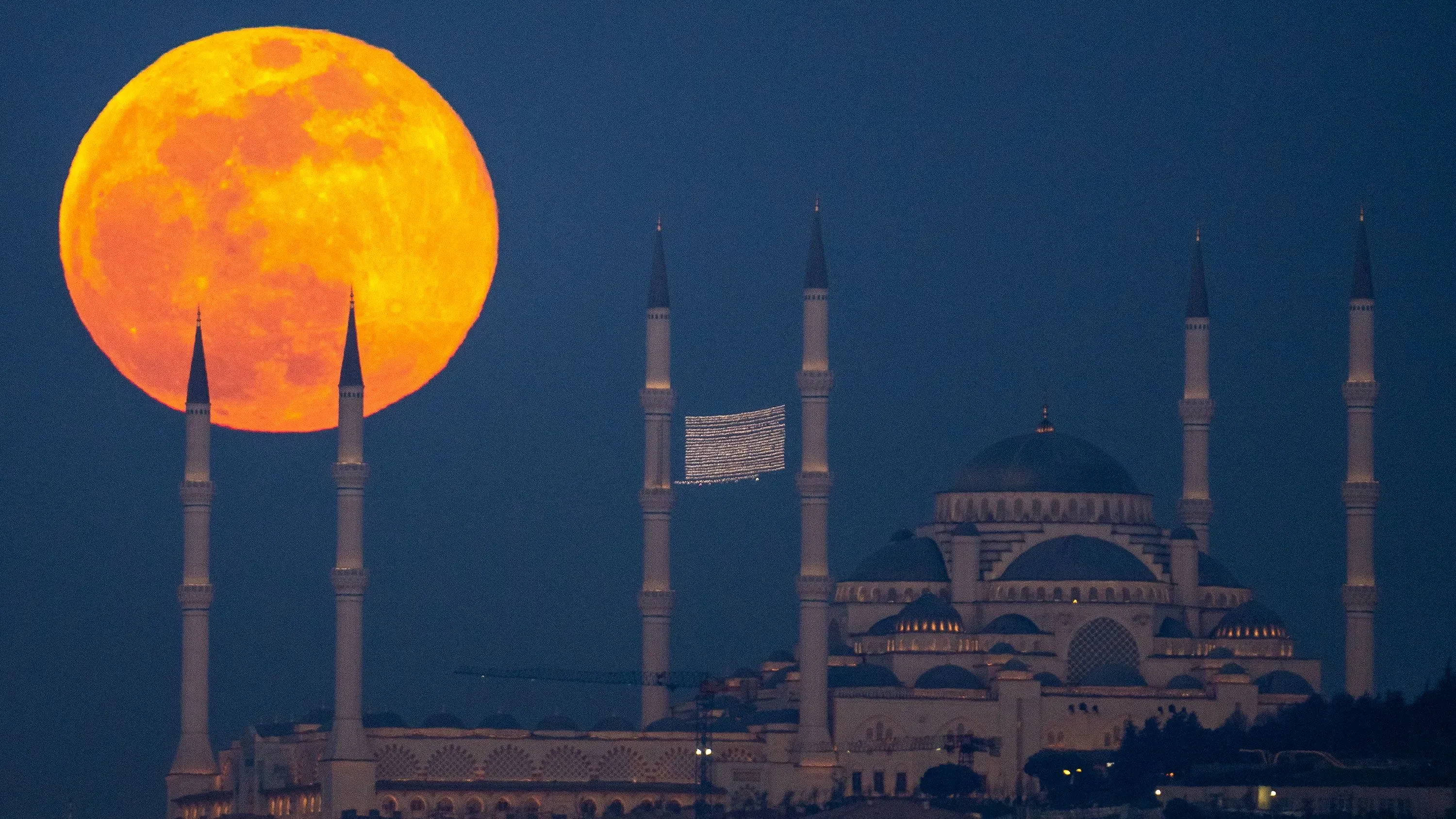 قمر الثلج المكتمل يضيء في السماء فوق مسجد، يظهر لونه البرتقالي المشرق مع مآذن بارزة في المقدمة.