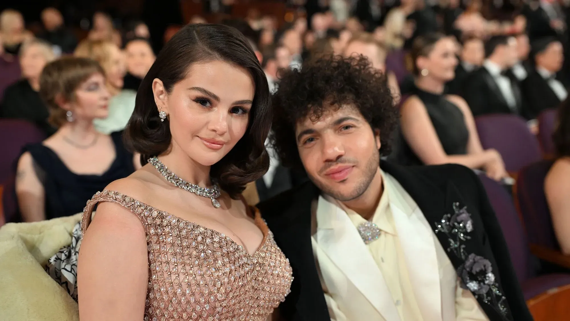 سيلينا غوميز وبيني بلانكو يجلسان معًا في حدث رسمي، حيث ترتدي غوميز فستانًا مزينًا، بينما يبدو الثنائي سعيدًا ومتماسكًا.