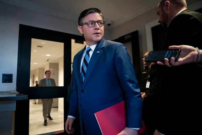 رجل يرتدي بدلة رسمية يحمل ملفًا، يخرج من غرفة اجتماعات في مبنى حكومي، مع وجود أشخاص آخرين في الخلفية، مما يعكس أجواء السياسة والقرارات المهمة.