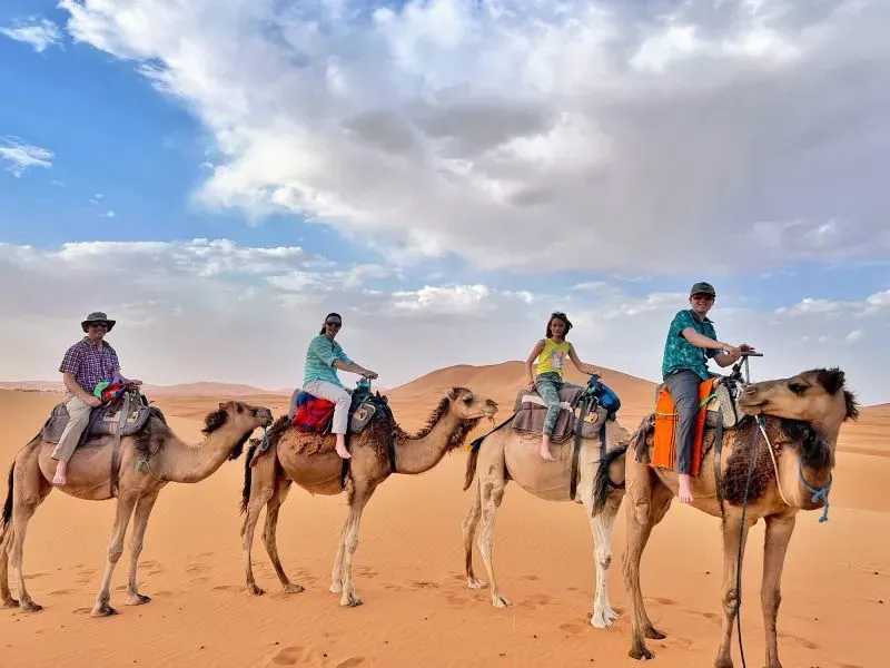 عائلة ألموند تستمتع بركوب الجمال في الصحراء، مع خلفية من الرمال والغيوم، أثناء مغامرتهم التعليمية حول العالم.