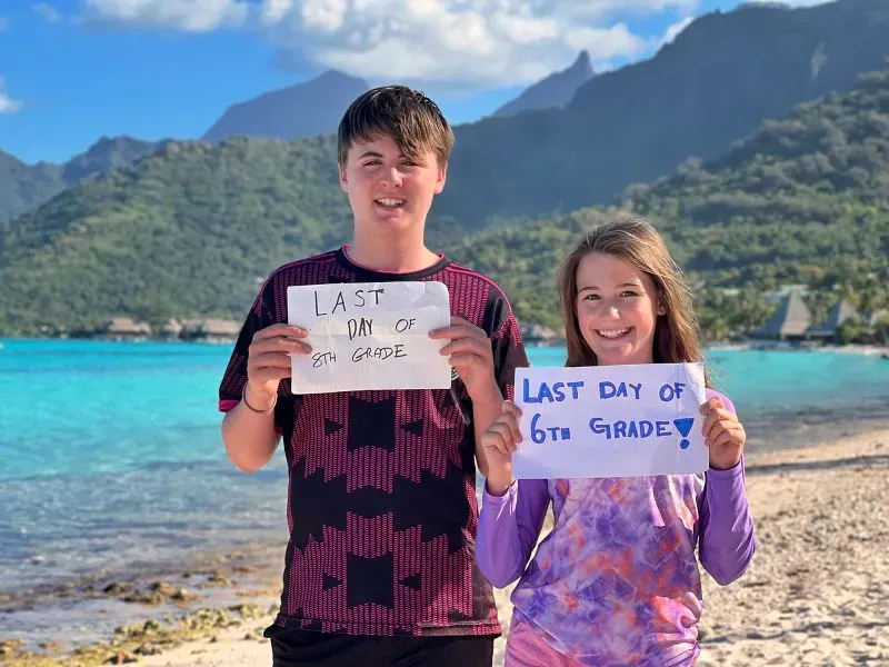 يحتفل طفلان على شاطئ بجزيرة استوائية، حيث يحملان لافتات مكتوب عليها "آخر يوم في الصف الثامن" و"آخر يوم في الصف السادس"، مع مناظر طبيعية خلابة خلفهما.