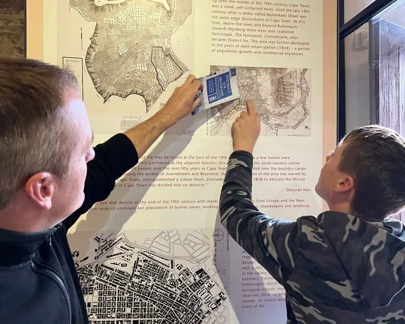 أب وابنه يتأملان خريطة تاريخية في كيب تاون، مستعرضين معلومات عن المدينة وتاريخها، أثناء رحلة تعليمية عائلية.