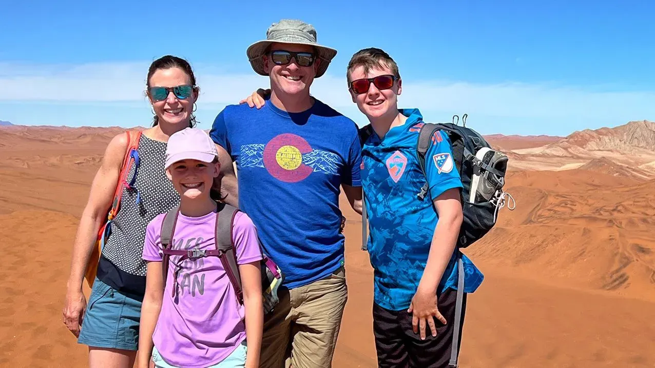 عائلة ألموند تتوسط منظرًا صحراويًا شاسعًا، حيث يظهر الوالدان وأطفالهما مبتسمين، مع خلفية من الرمال الحمراء والسماء الزرقاء.