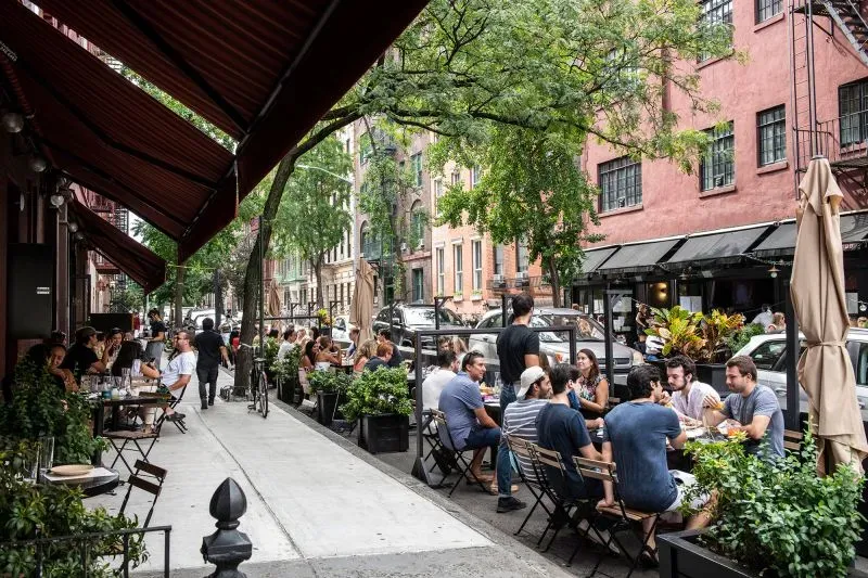 طاولات خارجية لمطعم في نيويورك، حيث يجلس الزبائن وسط أشجار ونباتات، مع أجواء حيوية تعكس تجربة تناول الطعام في الهواء الطلق.