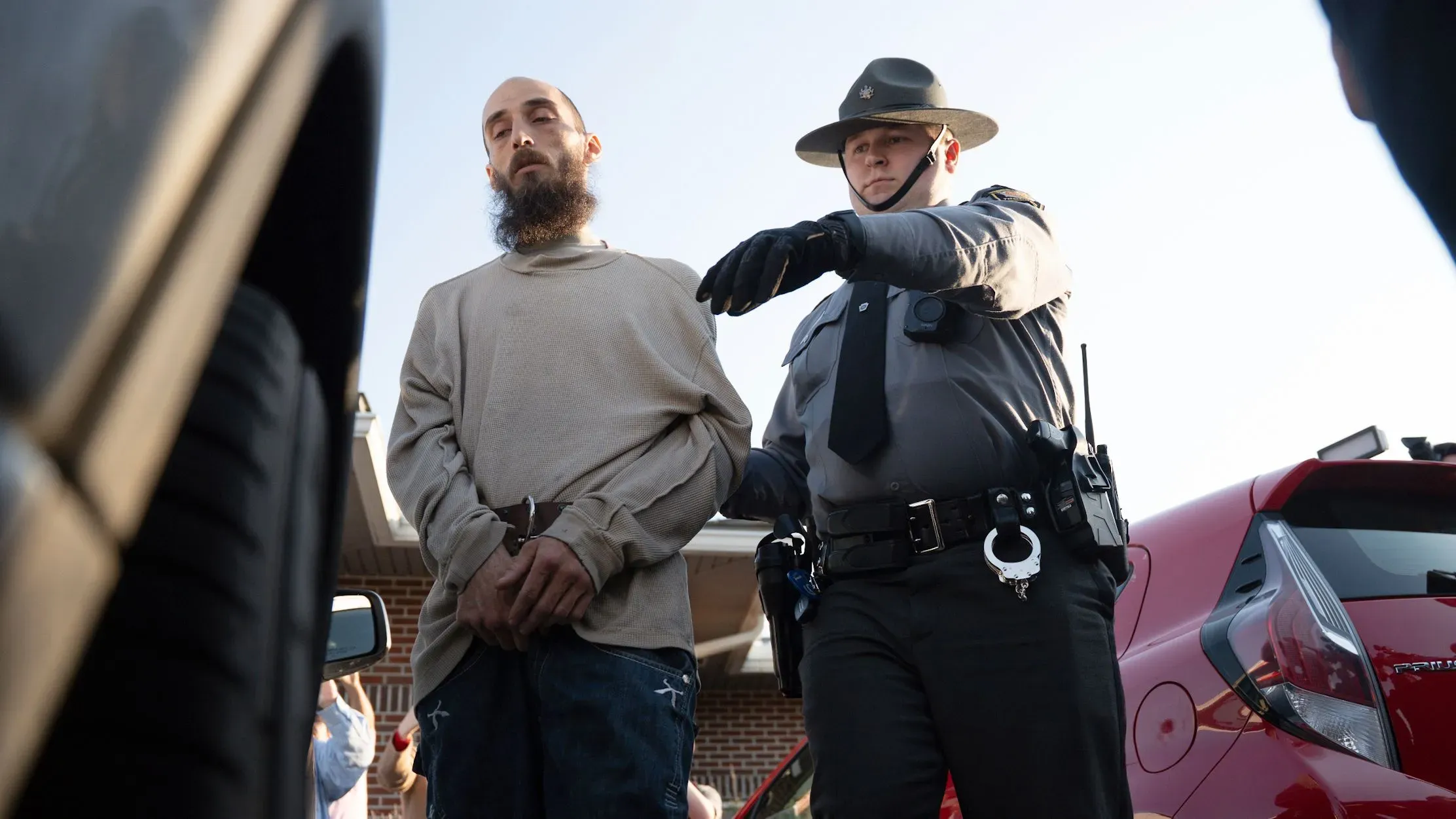 رجل مقيد اليدين يقف أمام ضابط شرطة، مع التركيز على التوتر في الموقف. الصورة تعكس تداعيات حادثة تتعلق بالصحة العقلية.