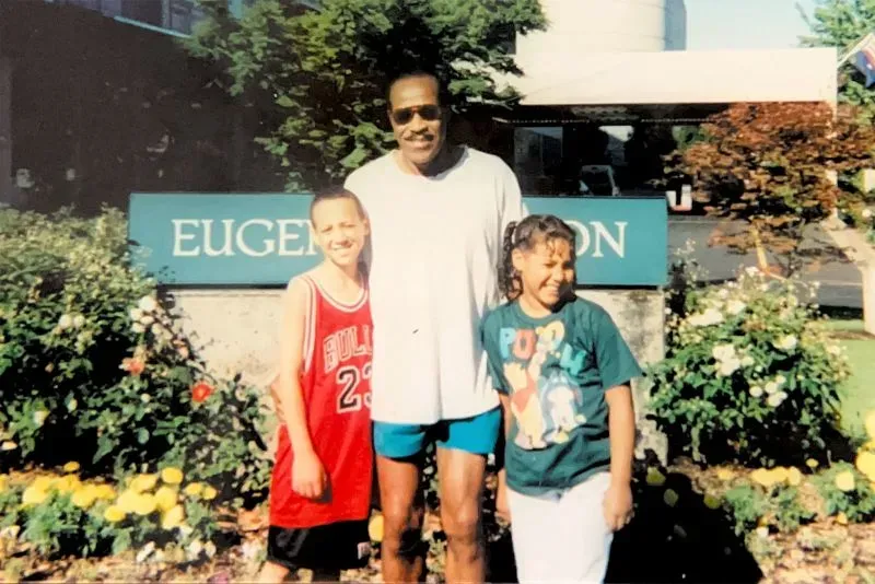 صورة تظهر رجلًا كبيرًا مع طفلين، أحدهما يرتدي قميص كرة سلة، أمام لافتة مكتوب عليها "Eugene" في حديقة مزهرة.