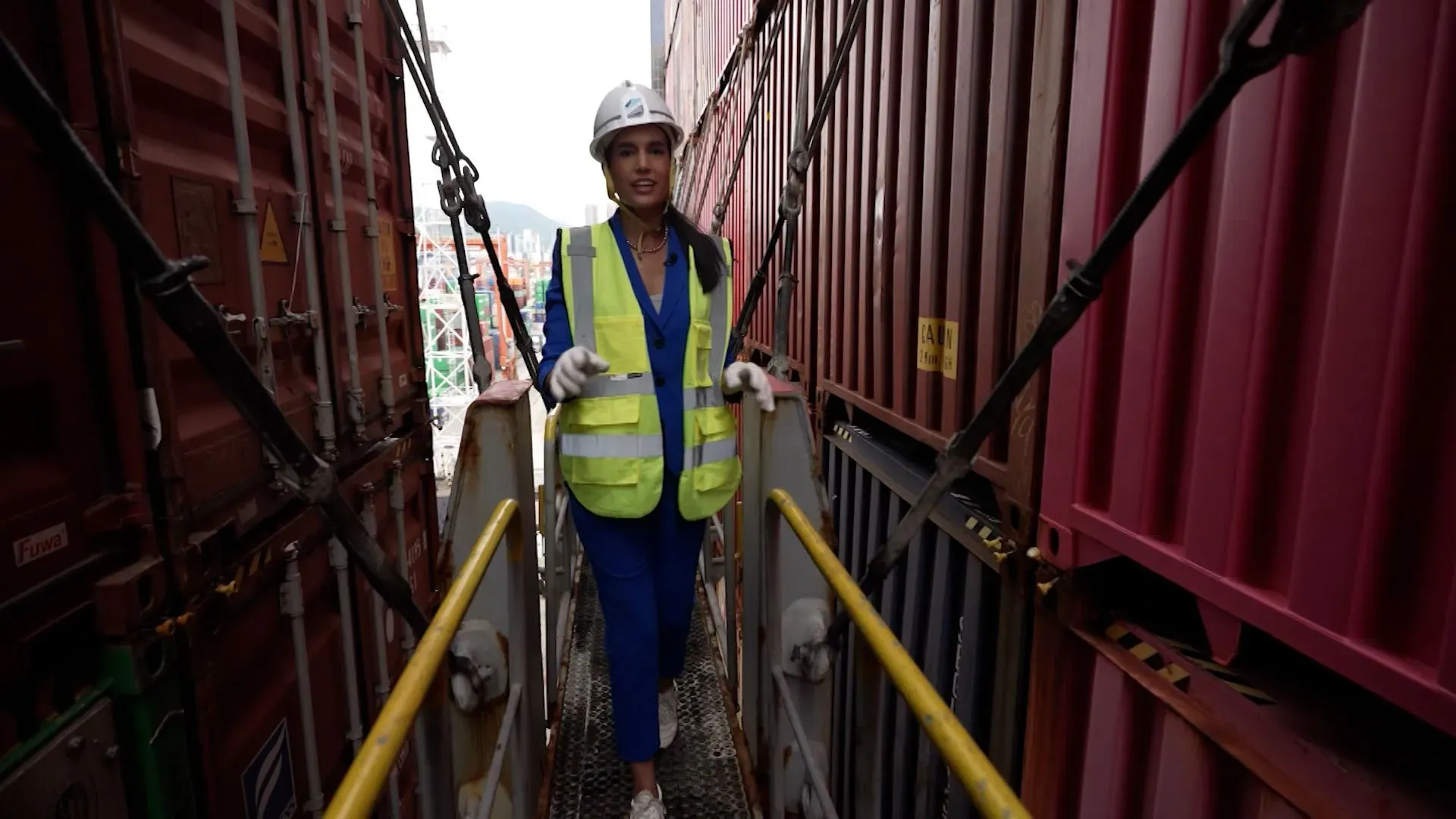 امرأة ترتدي سترة عاكسة وخوذة، تسير على ممر معدني بين حاويات شحن في ميناء هونغ كونغ، حيث يتم تفريغ السفينة ONE Modern.