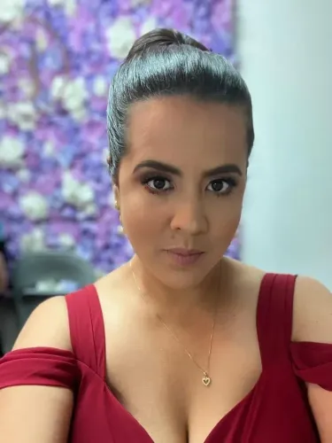 إستيفاني رودريغيز، صحفية كولومبية، تظهر في صورة سيلفي، ترتدي فستانًا أحمر، مع خلفية مزهرة. تغطي اعتقالات المهاجرين.