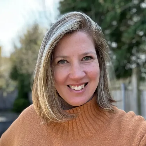 امرأة مبتسمة ترتدي سترة برتقالية، تظهر في خلفية طبيعية خضراء، تعكس الأمل والمرونة بعد حادث مأساوي.