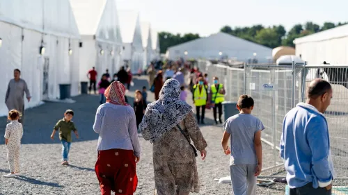 مجموعة من اللاجئين، بينهم نساء وأطفال، يسيرون في مخيم لمساعدتهم على التكيف في الولايات المتحدة، خلفهم خيام بيضاء وأفراد من طواقم الدعم.