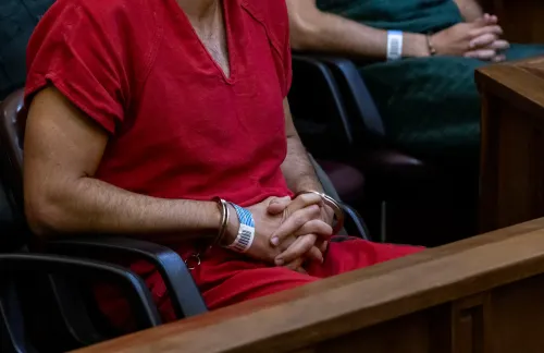 رجل يرتدي زي السجن الأحمر يجلس في قاعة المحكمة، مع يديه مقيدتين، بينما يتواجد آخرون في الخلفية. القضية تتعلق باتهامات بالاعتداء الجنسي.