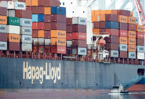 سفينة شحن محملة بالحاويات، تحمل شعار "Hapag-Lloyd"، ترسو في ميناء، مما يعكس النشاط التجاري وتأثير التعريفات الجمركية على التجارة.