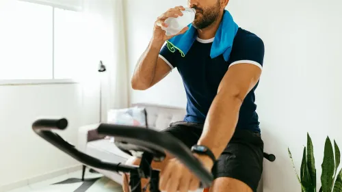 رجل يشرب الماء أثناء ركوبه على دراجة ثابتة في غرفة مضاءة جيدًا، مع مناشف حول عنقه ونباتات في الخلفية.