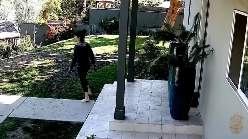 امرأة تسير في فناء منزلها، تحمل مسدسًا، بينما يظهر خلفها سياج خشبي وشرطتان في موقف متوتر قبل إطلاق النار.