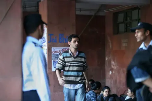 شاب يرتدي سترة مخططة يسير في ممر جامعة، بينما يظهر ضابط أمن وطلاب آخرون في الخلفية، مما يعكس أجواء الحرم الجامعي.