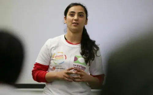 امرأة أفغانية تتحدث في حدث، ترتدي قميصاً يحمل شعارات حول حقوق الأطفال والمستقبل، تعبر عن أهمية كرة القدم للسيدات.