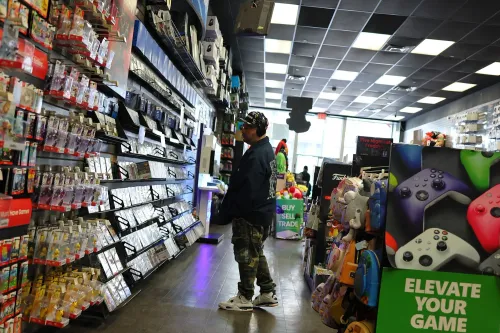 رجل يتصفح أرفف متجر GameStop المليئة بألعاب الفيديو والملحقات، مع وجود لافتة ترويجية تدعو لرفع مستوى تجربته في اللعب.