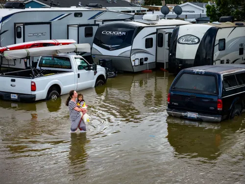 امرأة تحمل طفلاً في مياه الفيضانات، محاطة بمركبات غارقة، تعكس تأثير الأمطار الغزيرة والمد والجزر الملكي في شمال كاليفورنيا.