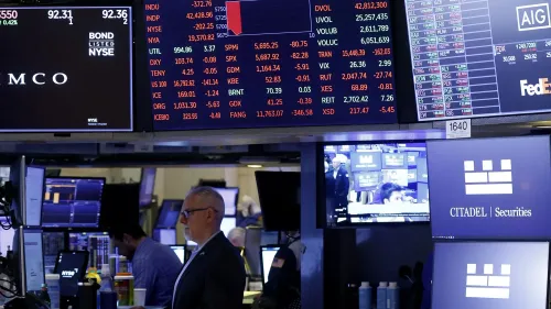 شاشة تداول في سوق الأسهم تعرض مؤشرات السوق وأسعار الأسهم، مع وجود متداولين في الخلفية، تعكس حالة عدم اليقين الاقتصادي.