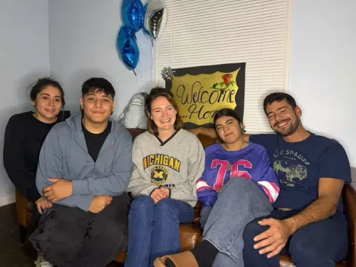 مجموعة من الطلاب مع معلمتهم في منزلهم بعد لم الشمل، مع لافتة ترحيب خلفهم، تعبير عن الفرح والأمل بعد فترة من الاحتجاز.