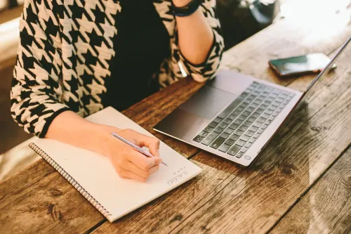 امرأة تجلس أمام جهاز كمبيوتر محمول، تكتب في دفتر ملاحظات على طاولة خشبية، مع وجود هاتف محمول بجانبها، تعكس مشاعر التخطيط والتركيز.