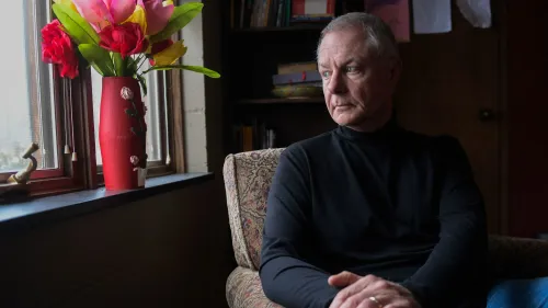 رجل مسن يجلس بجوار نافذة مضاءة، يتأمل بجدية، مع زهور ملونة في مزهرية حمراء بجانبه، مما يعكس أجواء من التأمل والقلق.