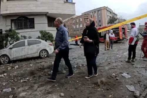 مشهد لعدد من الأشخاص يسيرون في منطقة متضررة في طهران، مع وجود سيارات إسعاف وخطوط تحذير، مما يعكس آثار الهجمات على البنية التحتية.