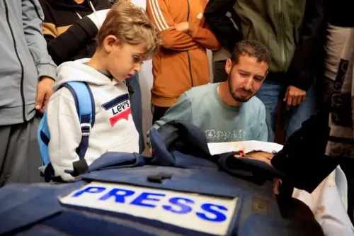 مشهد مؤثر يظهر طفلًا ينظر بحزن إلى صحفي ميت، بينما يتجمع حوله أشخاص آخرون، مع التركيز على سترة مكتوب عليها "PRESS".