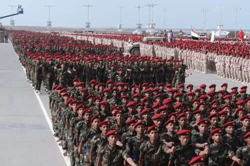 جنود يرتدون زيًا عسكريًا وخوذات حمراء، يسيرون في صفوف منتظمة خلال عرض عسكري، مما يعكس أهمية القوة العسكرية في المنطقة.