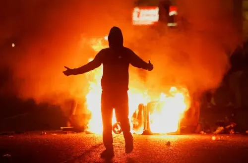شخص ملثم يقف أمام حريق في الشارع خلال أعمال شغب في أيرلندا الشمالية، حيث تندلع الاضطرابات بسبب اعتداء جنسي مزعوم.